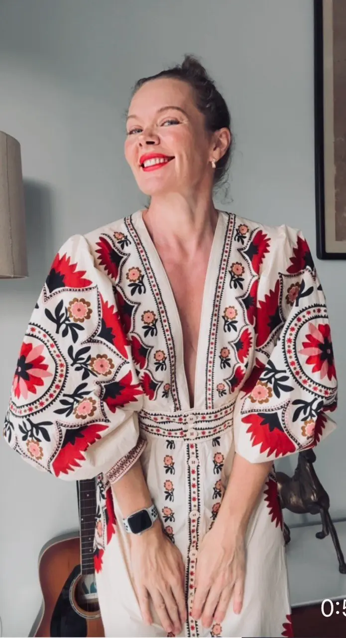 A woman in a Ukrainian embroidered costume smiles and stands next to a guitar. She is wearing a dress with bright red and black patterns on a white background, which gives the image a national flavor. The woman looks away, a happy expression on her face. Her hair is pulled back, and a wearable device, possibly a smartwatch, is visible on her wrist.